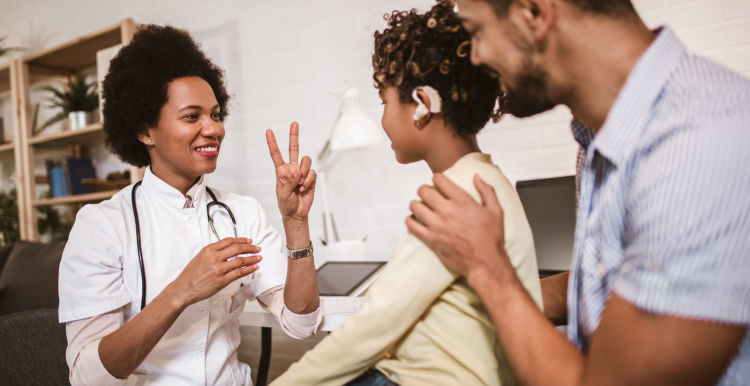 In a GP office. The GP has a stethoscope around her neck. She is communicating with a young patient in British Sign Language. The young patient has an audio prothesis. His Dad is behind him, a hand on his shoulder. The three of them are smiling. 