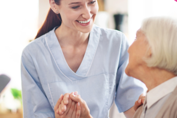 A nurse wearing a blue uniform is smiling warmly at an elderly patient. The patient is seated. They are holding hands.