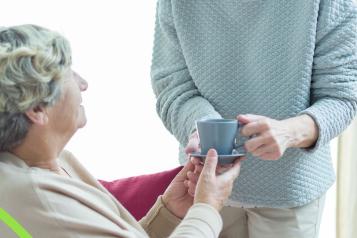 This is a photo of two women. One elderly woman is seated on a sofa. The other woman is standing next to her, giving her a cup of tea while smiling warmly.
