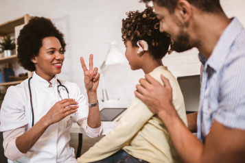 In a GP office. The GP has a stethoscope around her neck. She is communicating with a young patient in British Sign Language. The young patient has an audio prothesis. His Dad is behind him, a hand on his shoulder. The three of them are smiling. 