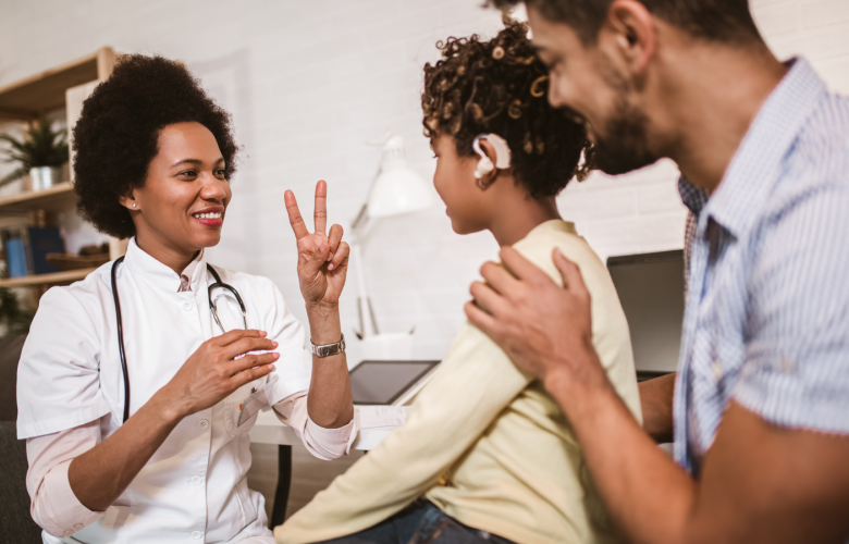 In a GP office. The GP has a stethoscope around her neck. She is communicating with a young patient in British Sign Language. The young patient has an audio prothesis. His Dad is behind him, a hand on his shoulder. The three of them are smiling. 