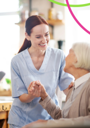 A nurse wearing a blue uniform is smiling warmly at an elderly patient. The patient is seated. They are holding hands.