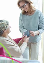 This is a photo of two women. One elderly woman is seated on a sofa. The other woman is standing next to her, giving her a cup of tea while smiling warmly.